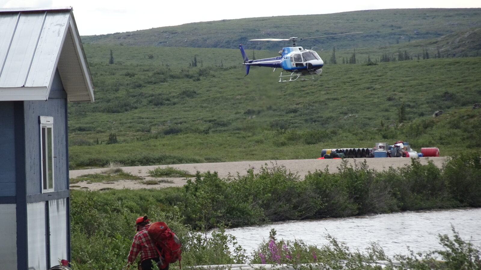 A geologist is walking towards a helicopter hovering above ground in field. Drums on ground.