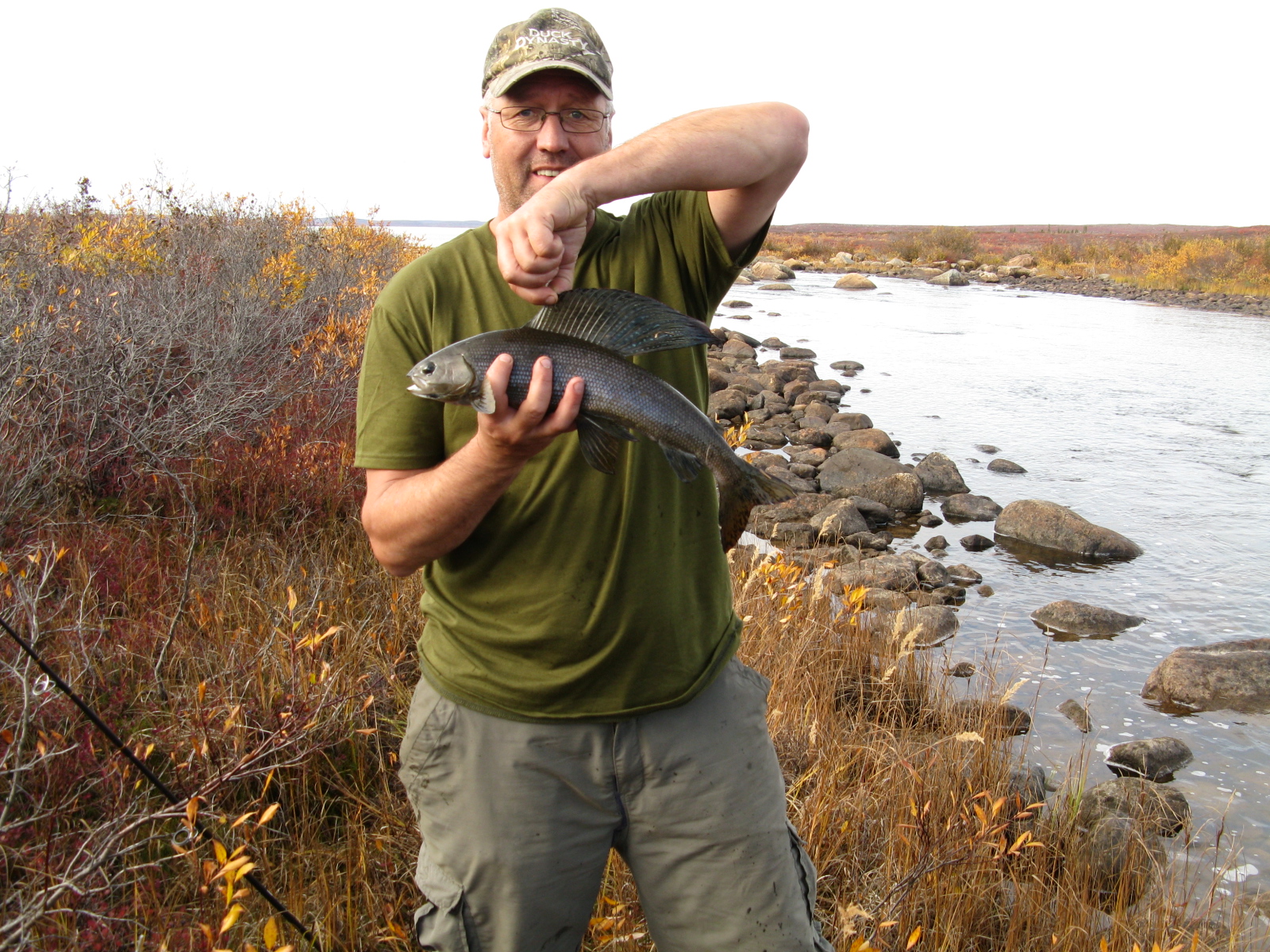 A man smiling, holding the dorsal fin of an Arctic Grayling on the shore of the Itchen River