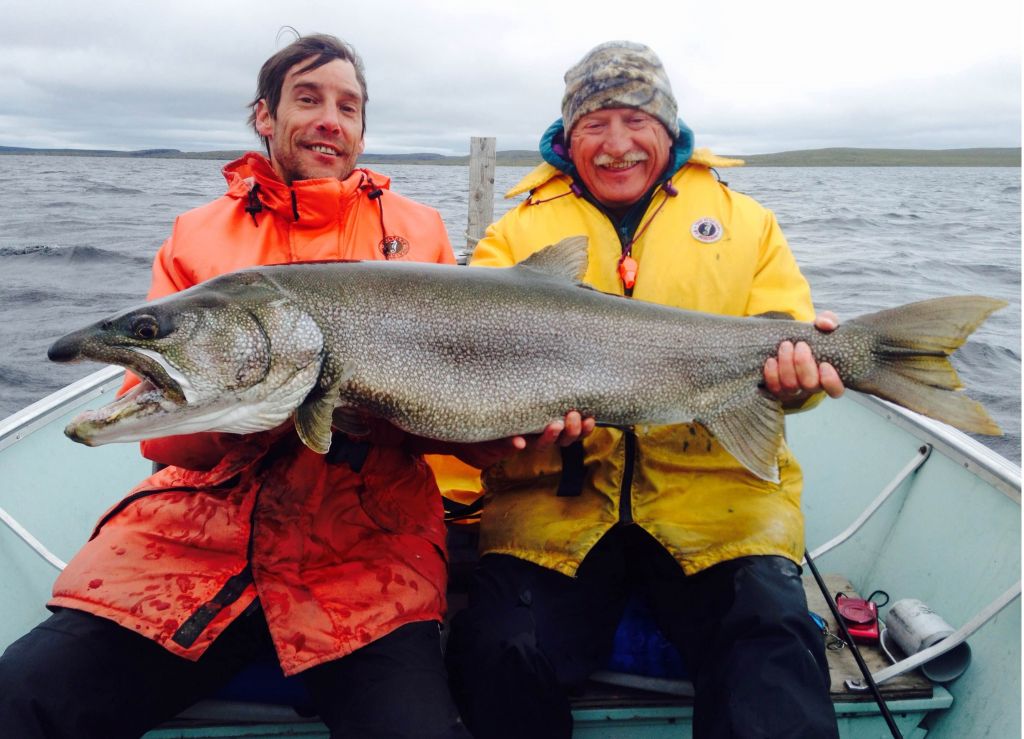 Two men wearing life jackets in boat smiling while holding a trophy lake trout. Passion angling supersedes comfort.