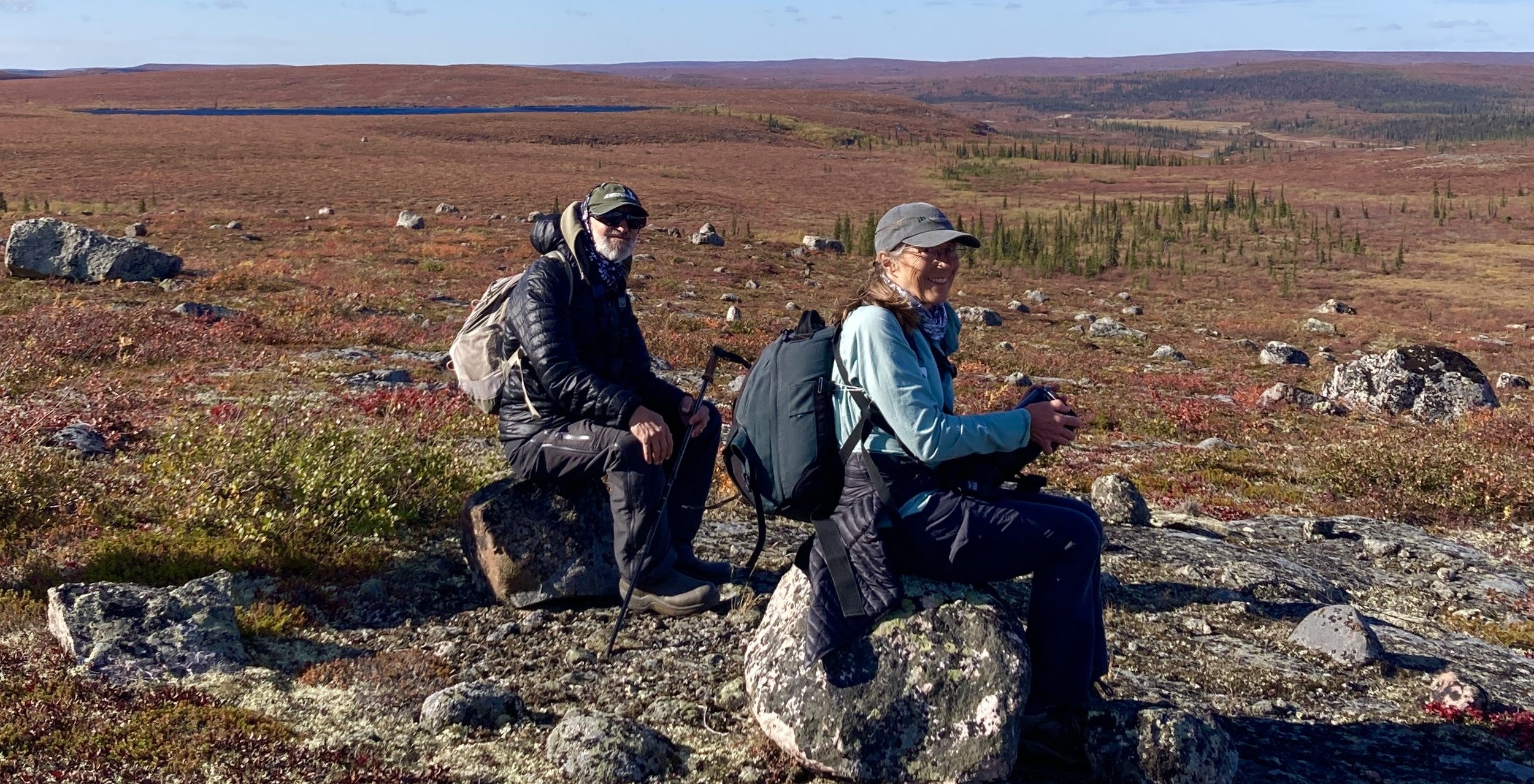 Two people sitting on two boulders with camera in hand overlooking incredible fall barrenland scenery