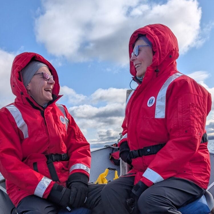 Two smiling guests wearing red hooded flotation jackets, gloves, and sunglasses sharing a laugh aboard a tour boat on Point Lake, enjoying an outdoor expedition in the Canadian Barrenlands with Peterson’s Point Lake Lodge, Northwest Territories