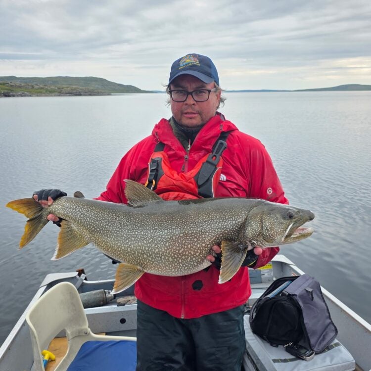 An angler wearing glasses, a navy Peterson’s Point Lake Lodge cap, and a bright red waterproof jacket proudly displays a massive trophy lake trout aboard a boat on the calm waters of Point Lake during a Canada lake trout fishing excursion in the Northwest Territories.