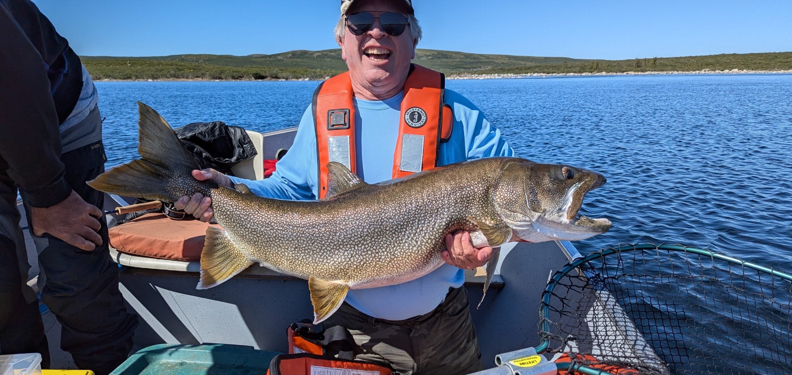 A smiling fisherman in an orange life vest holding a large Lake Trout on a boat at Peterson’s Point Lake Lodge under a clear blue sky.