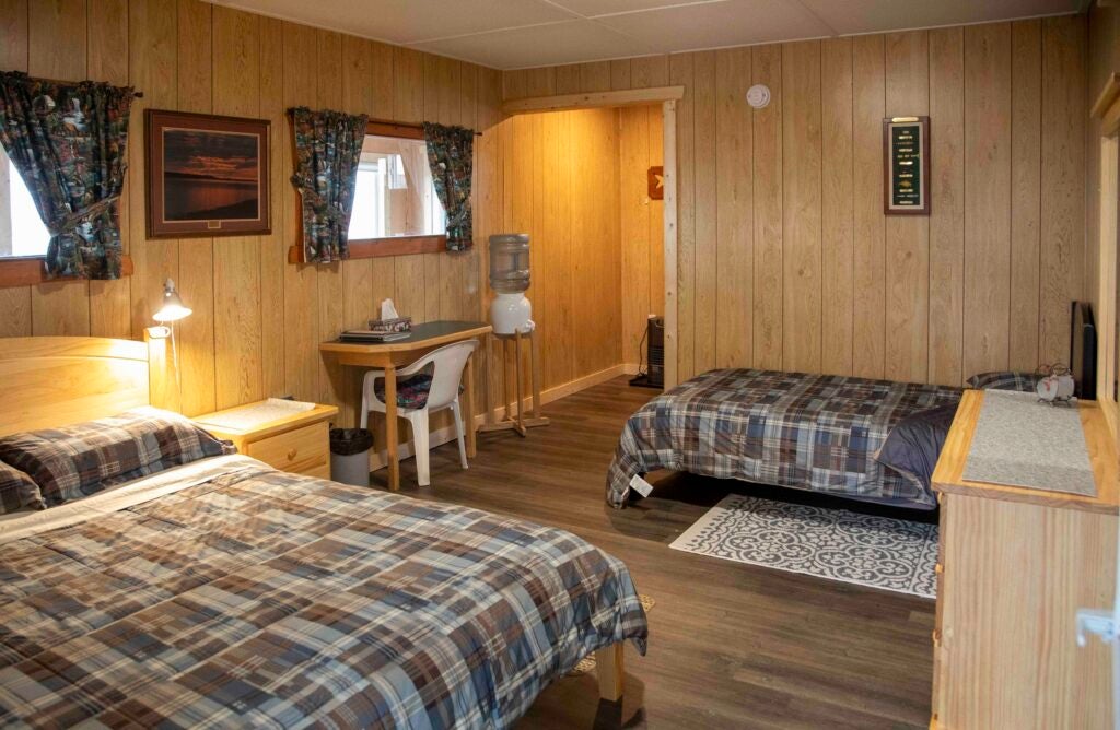 Interior of cabin suite featuring wood-paneled walls. The room includes a queen sized bed and a twin bed, both with blue and brown plaid bedding. A small wooden desk with a chair, a water cooler, and a dresser complete the space, illuminated by warm bedside lighting.
