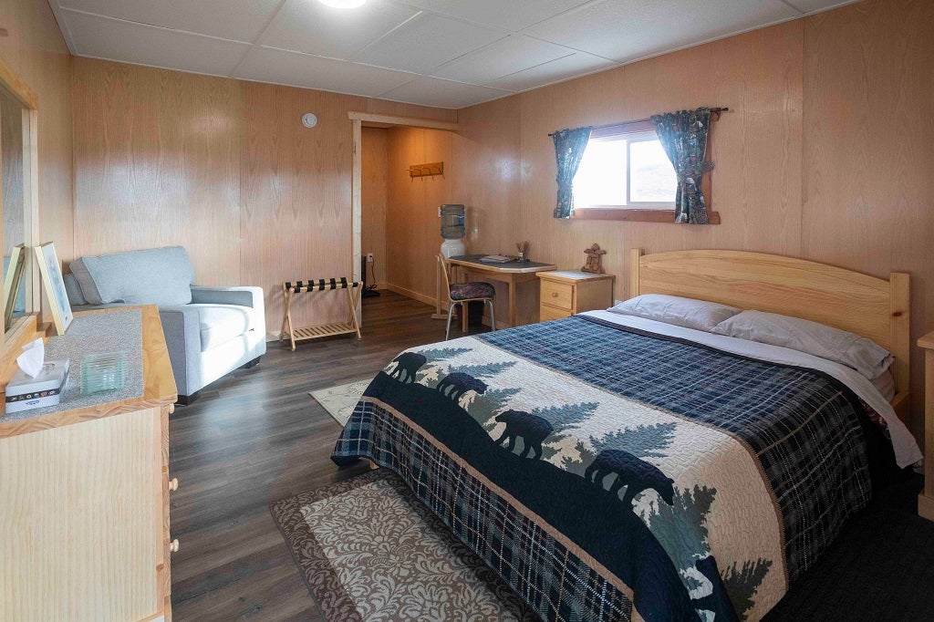 Interior of a cozy cabin showing a queen bed topped with a decorative quilt featuring a silhouette bear and pine tree pattern. The room includes a comfortable grey armchair, a small work desk, dresser, a luggage rack, and large windows with patterned curtains, creating a bright and inviting atmosphere.