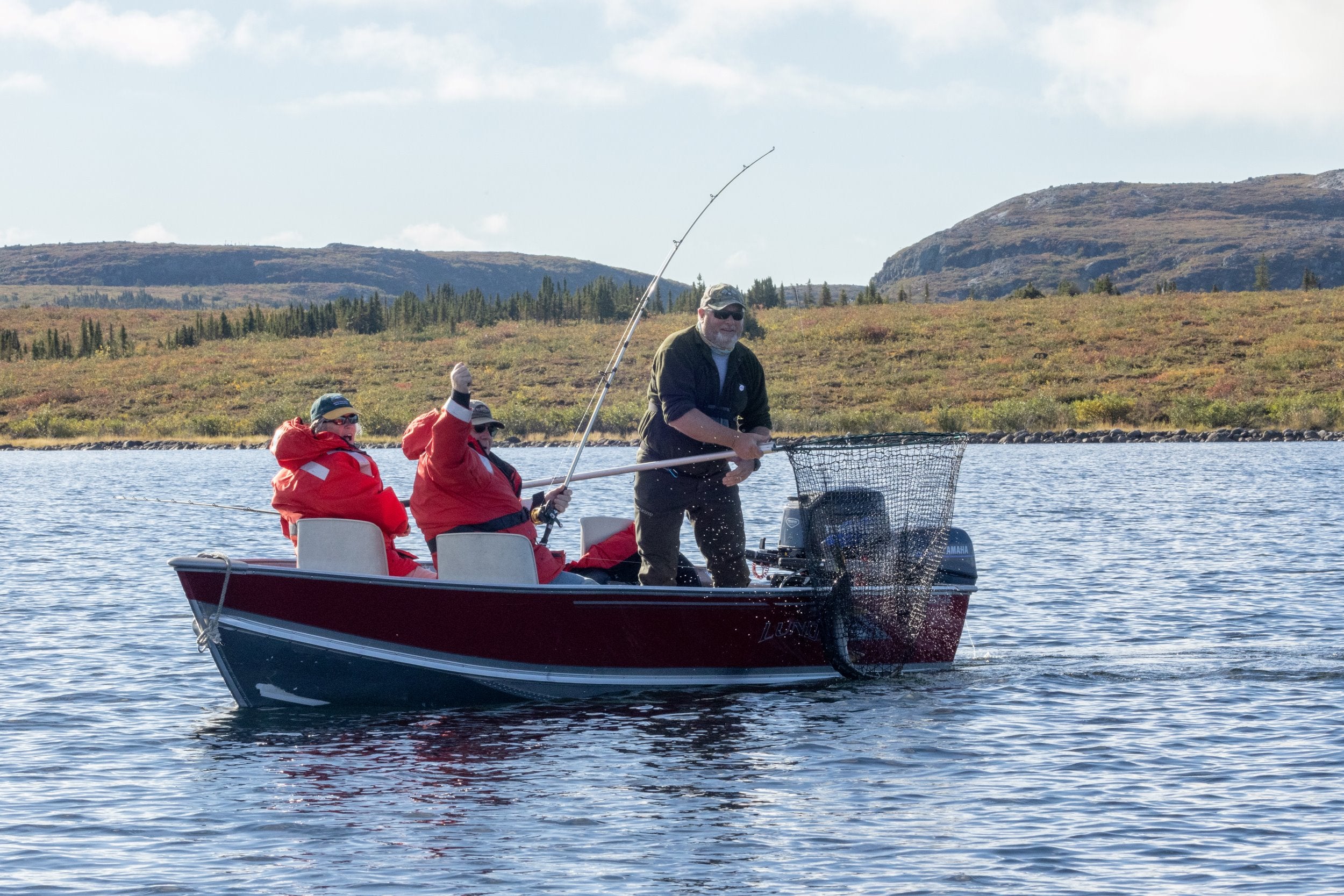 A boat with two anglers and a guide on a remote lake in the barrens. One guest excitedly waves his arm in celebration as the guide nets a freshly caught lake trout.