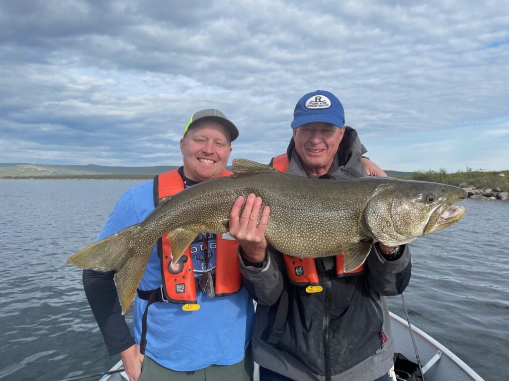 Two men with big smiles, one holding a large lake trout standing beside his son before releasing it into the water.