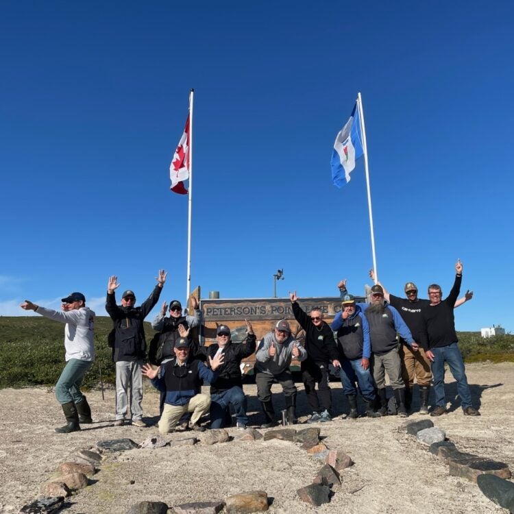 Group with hands in the air celebrating a terrific fishing trip, blue skies with Petersons Point Lake Lodge sign in the background with Canadian and Northwest Territories Flags flying.