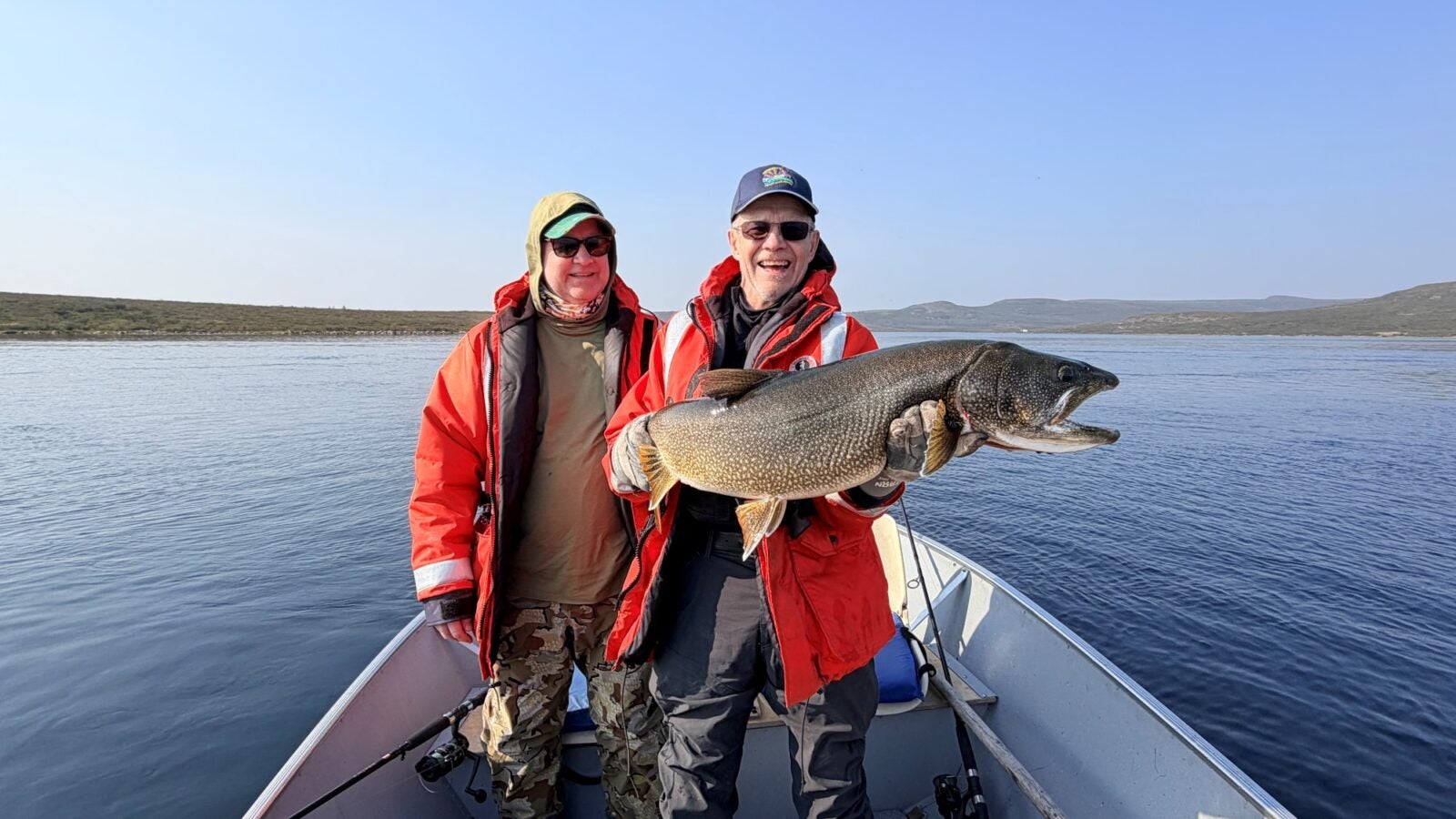 Two fishing partners wearing Mustang floater coats aboard a boat, holding up a massive trophy lake trout caught during a Canada lake trout fishing trip on the open waters of Point Lake, Northwest Territories.