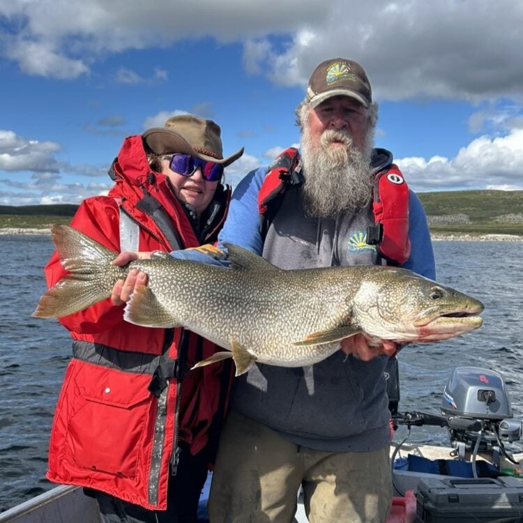 A smiling female angler in a red jacket and brimmed hat, alongside a bearded guide wearing a Peterson’s Point Lake Lodge cap, proudly holding a giant trophy lake trout aboard a boat on Point Lake during a Canada lake trout fishing adventure in the Northwest Territories.